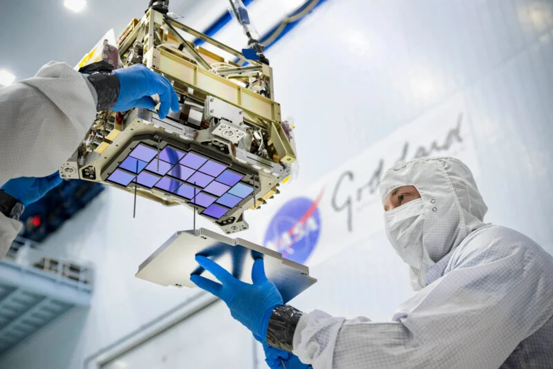 航天图片资料 · A technician in a cleanroom suit and blue gloves assembles a space instrument with a grid of purple sensors.