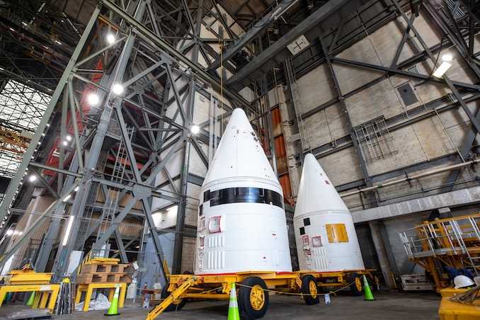 航天图片资料 · Two large white rocket-shaped payload fairings sit on yellow transport carts inside a tall industrial facility. The building’s interior is filled with steel beams, platforms, and bright overhead lights.