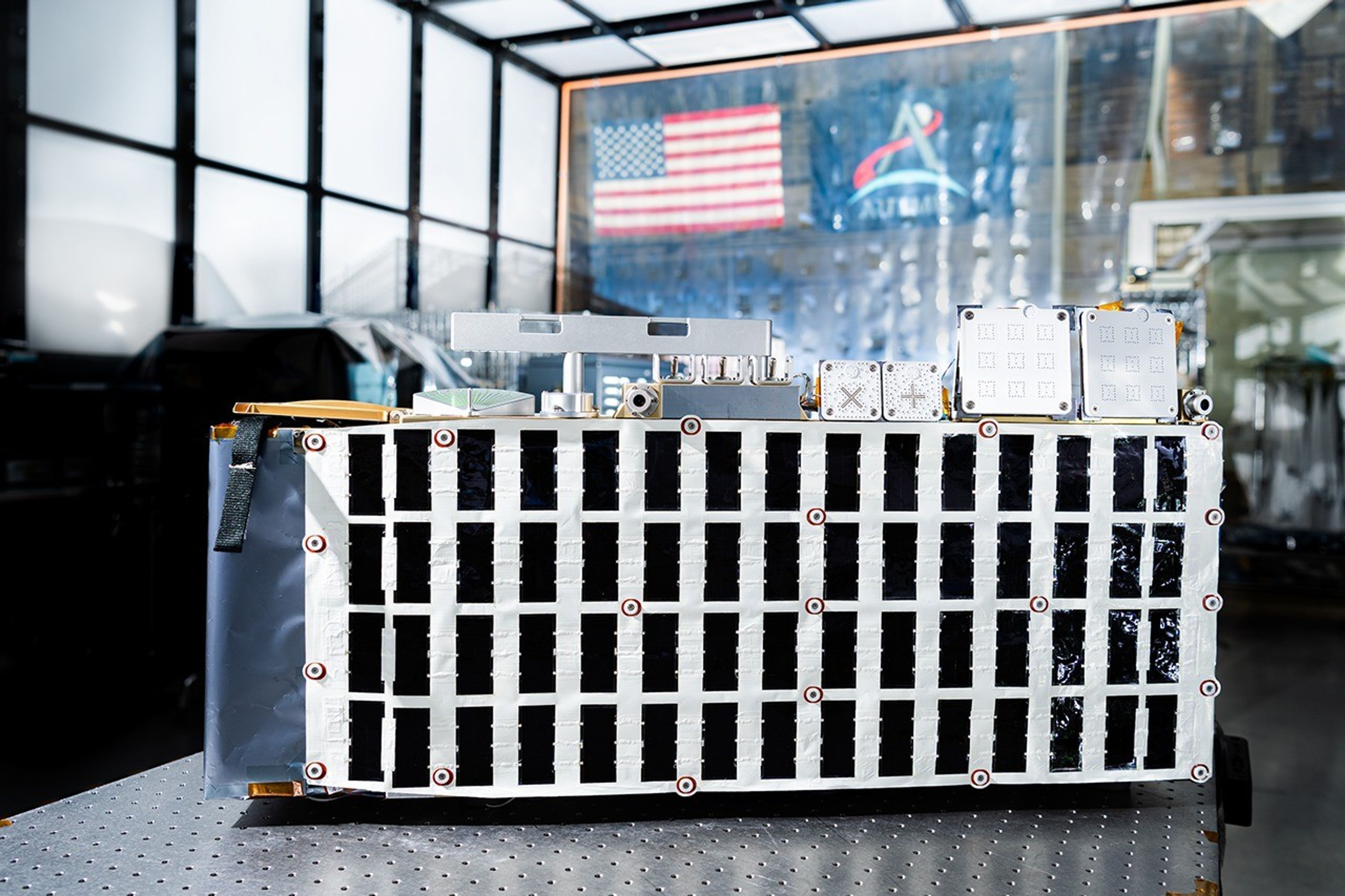 航天图片资料 · Rectangular box-shaped device resting on a metal table. The front face is covered with a grid of many small black rectangular panels bordered in white. Metal components, brackets, and small box units are mounted along the top. The background shows a large windowed wall with a blurred American flag and an Artemis flag visible behind the device.