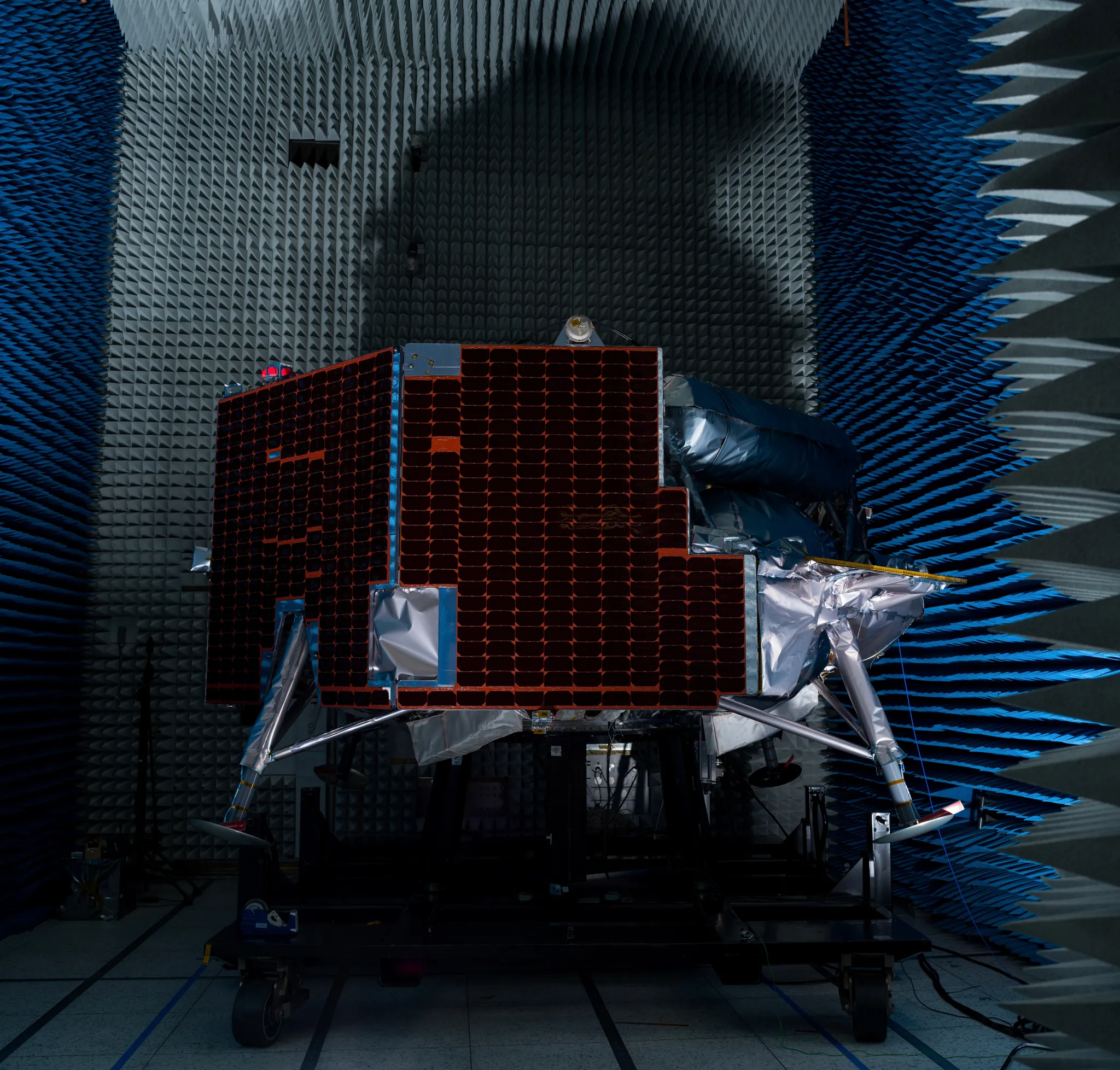 航天图片资料 · A lunar lander equipped with dark solar panels and foil-wrapped legs sits on a platform inside a dark anechoic chamber lined with blue and gray foam spikes.
