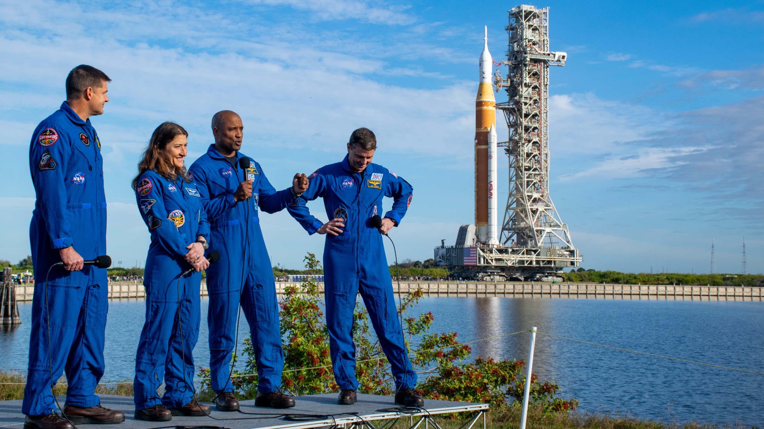 航天图片资料 · Four humans in blue jumpsuits stand outside on a raised platform. A big orange rocket and launcher tower can be seen in the background.