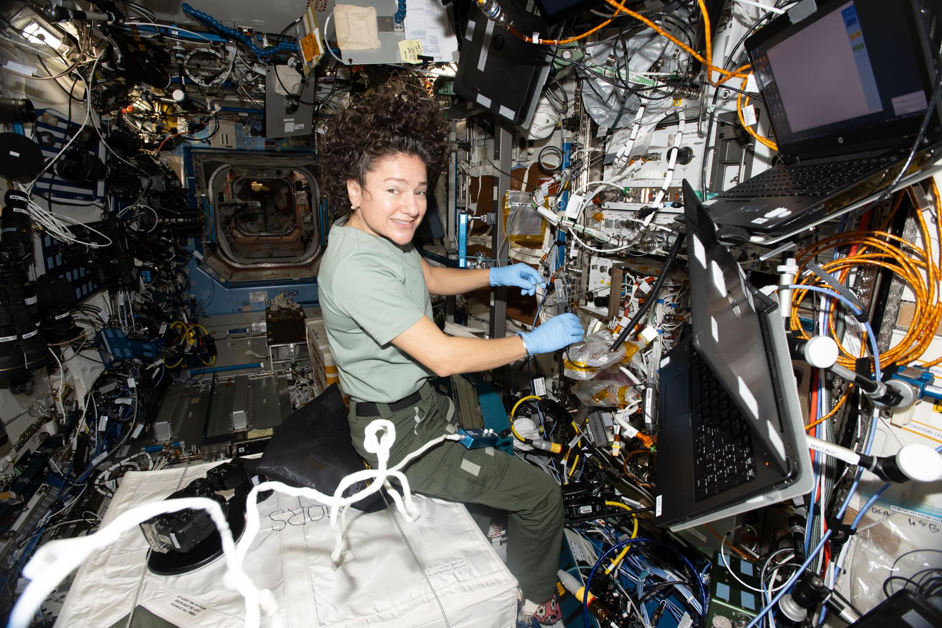 航天图片资料 · NASA astronaut Jessica Meir wears blue gloves and smiles at the camera while working with equipment aboard the International Space Station. Cables, tubes, and hardware cover the wall around her, and two laptops are mounted to her right. She rests on a large white padded container, giving her a seated appearance in microgravity.