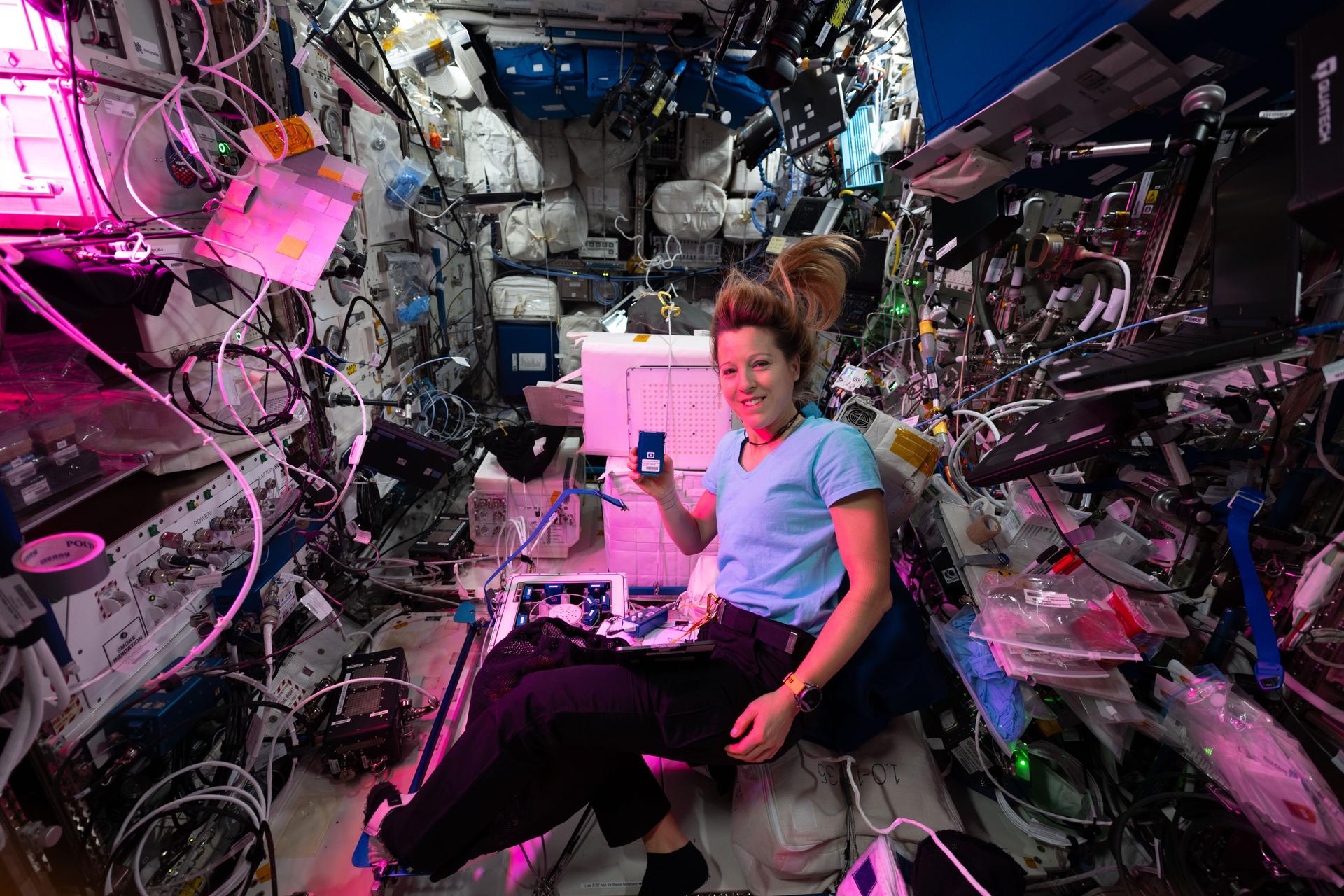 航天图片资料 · ESA (European Space Agency) astronaut Sophie Adenot floats on the International Space Station, surrounded by wires, compartments, and plastic packages. There is a pink light emanating from the left side of the image and it shines on Sophie and the objects around her. She smiles and holds up a blue, metallic rectangle in her hand.