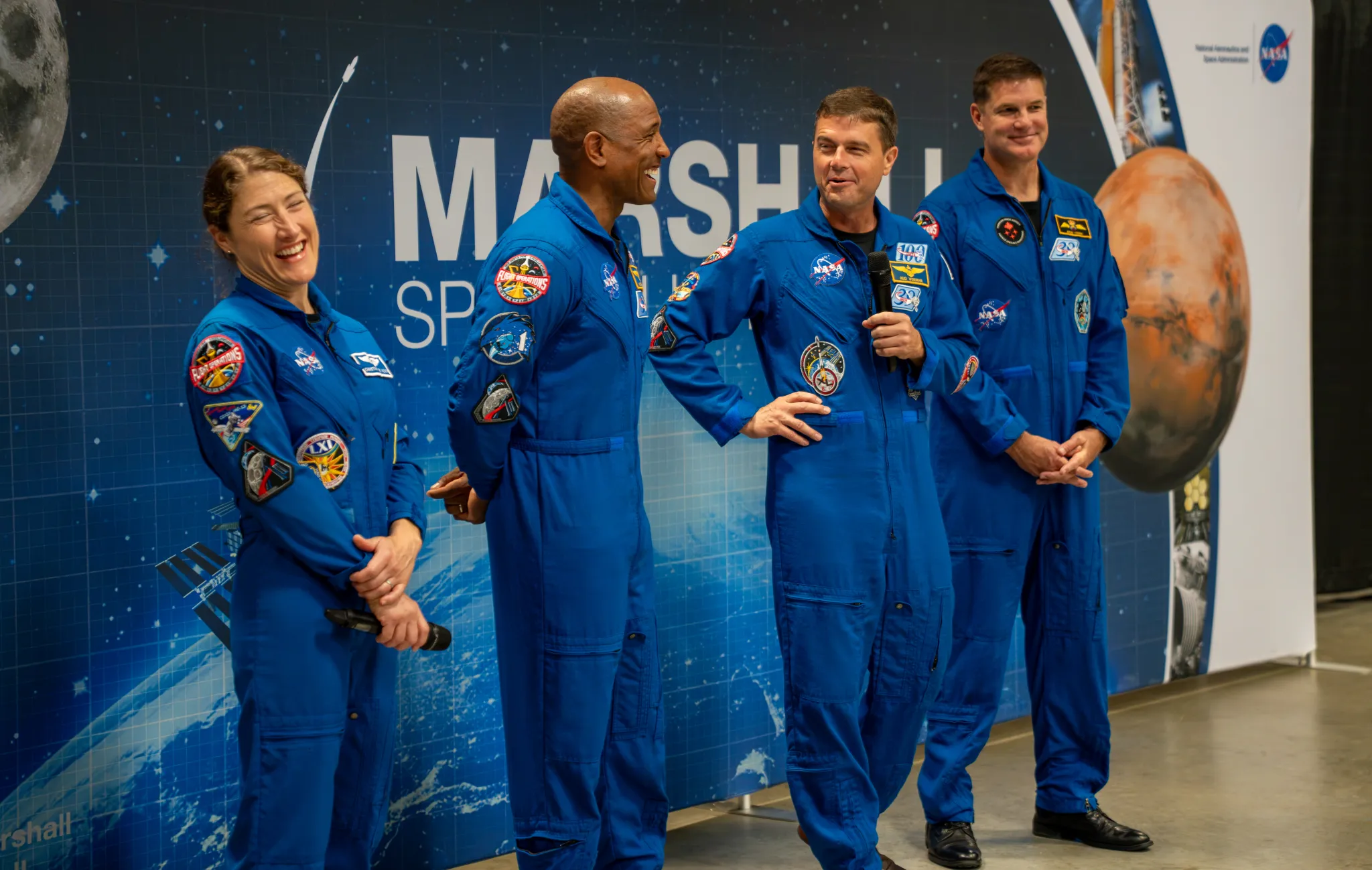 航天图片资料 · The Artemis II crew smile and laugh. They are wearing blue jumpsuits with patches on them. Christina Koch, left, and Reid Wiseman, second from right, are holding microphones.