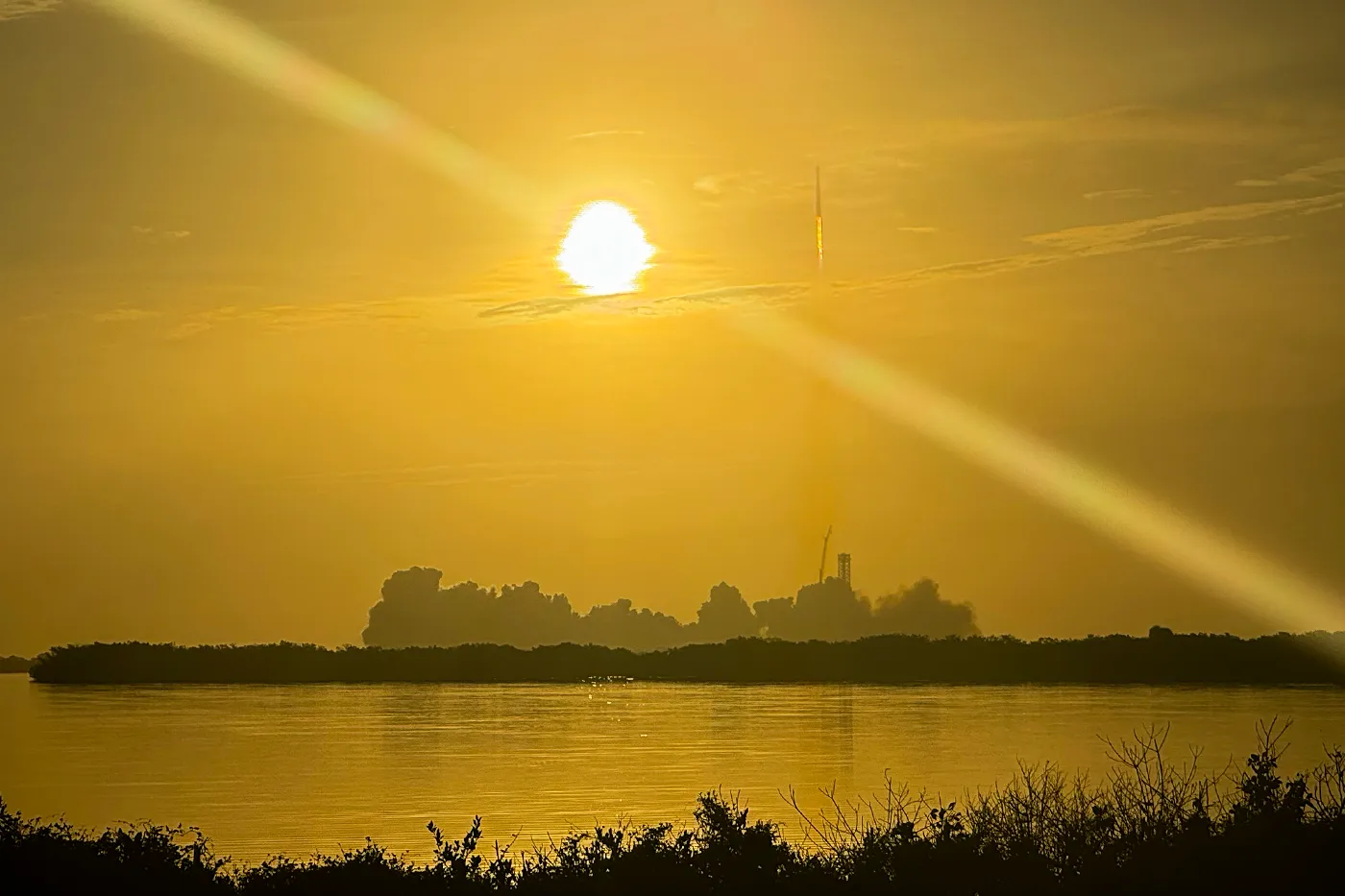 航天图片资料 · A rocket standing on its launch pad near the horizon at sunrise, seen across water with a bright sun overhead.