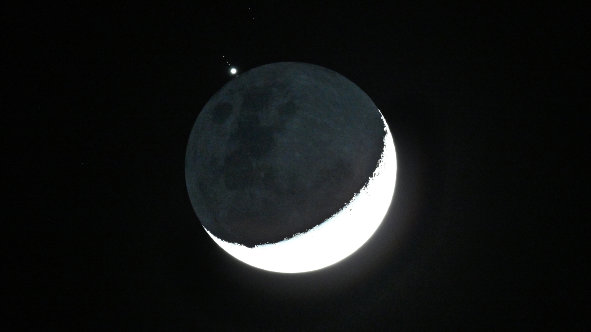 航天图片资料 · The crescent moon is shown in a black night sky with a bright point of light representing Jupiter positioned above its shadowed surface, with small star-like objects representing its largest moons.