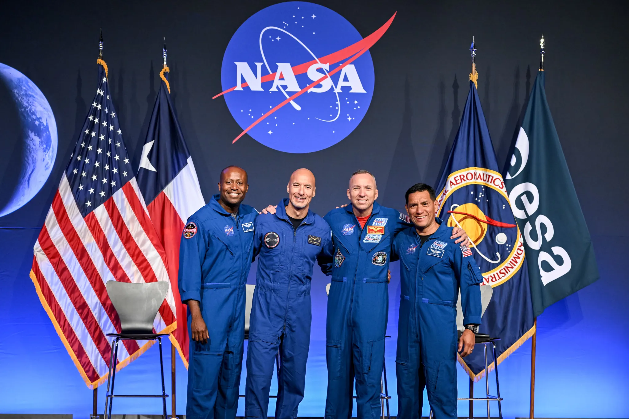 航天图片资料 · From left to right, NASA astronaut Andre Douglas, ESA (European Space Agency) astronaut Luca Parmitano, and NASA astronauts Randy Bresnik and Frank Rubio take a photo together. Their arms are linked around each other's shoulders. Behind them are the flags of the United States, Texas, NASA, and ESA.