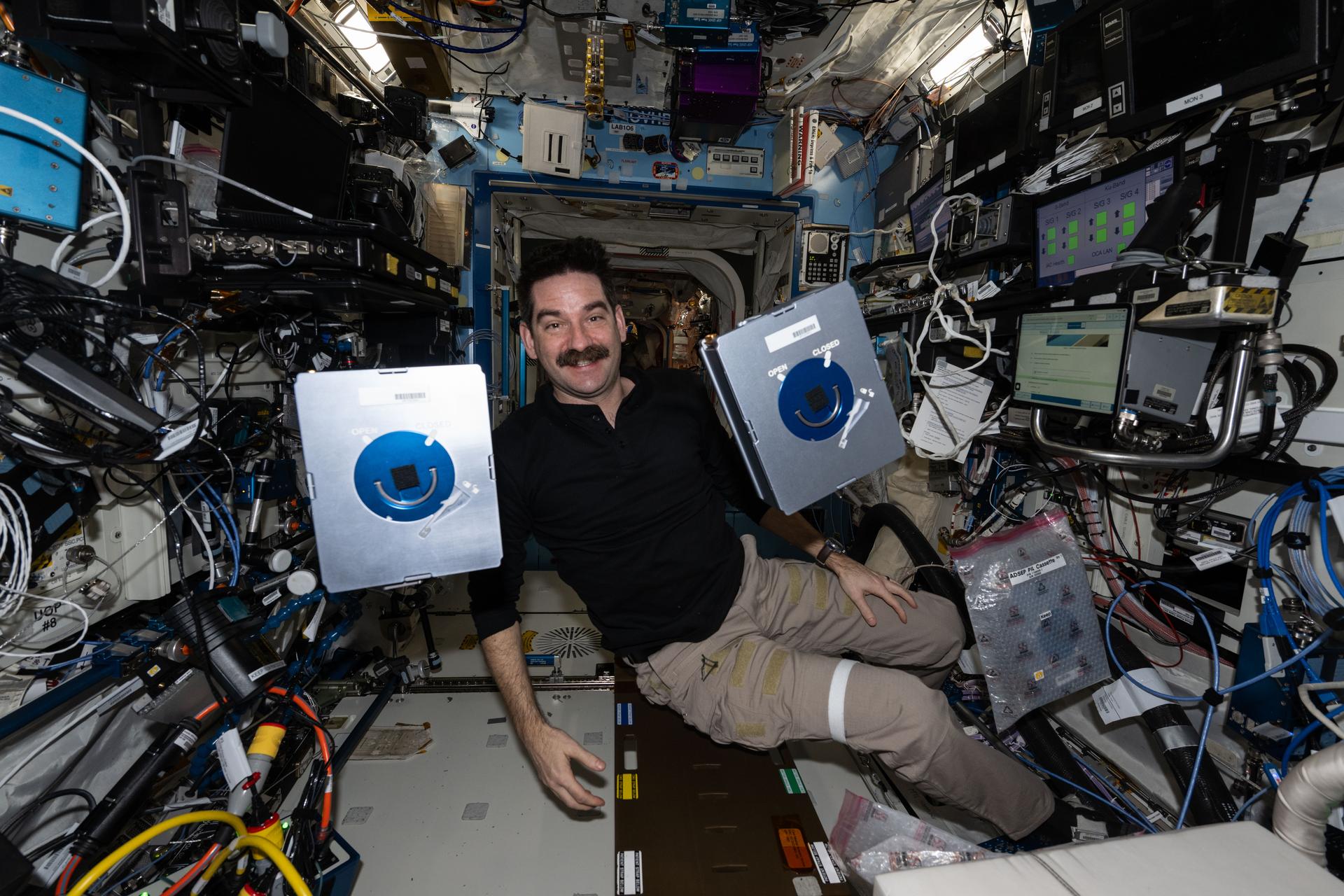 航天图片资料 · NASA astronaut Jack Hathaway floats and smiles at the camera, his face framed by two metallic silver boxes. Around him, the International Space Station’s walls are densely packed with wires, tubes, computer screens, and other hardware.
