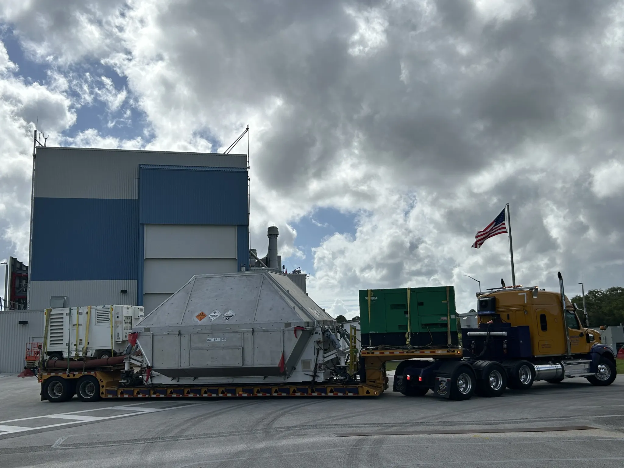 航天图片资料 · A large truck with a yellow cab hauls a wide, metal, container-like structure on a low trailer, with additional equipment secured behind it. An American flag flies from the truck as it sits on a paved area beside a tall industrial building. Thick, backlit clouds fill the sky above at NASA's Kennedy Space Center in Florida on Tuesday, April 28, 2026. Photo credit: NASA/Tiffany Fairley